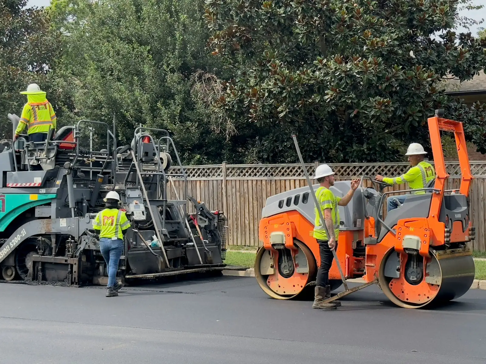 A paving crew in high visibility vests working with a paver and roller on a suburban street